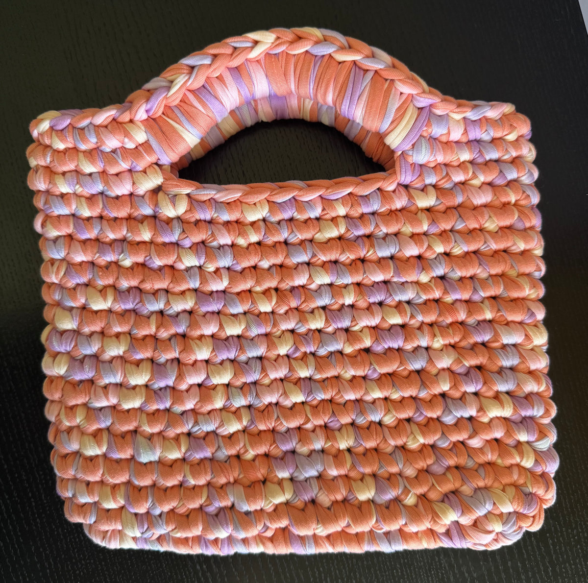 A woven tote bag made from variegated orange, pink, purple, and white yarn, featuring a single integrated handle, set against a dark textured background.